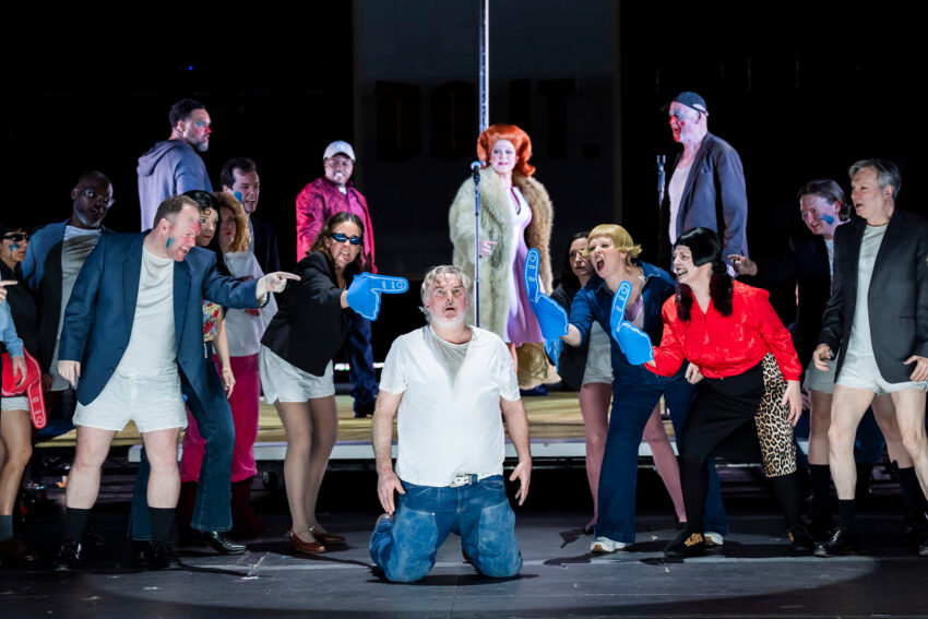 A man kneels centre stage in a white T-shirt and underwear, surrounded by a diverse group of actors in colourful, eccentric costumes. Many point at him—some with blue foam fingers—evoking the dramatic spectacle of Rise and Fall of the City of Mahagonny.