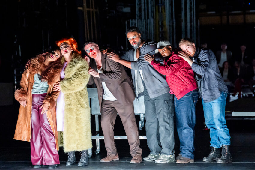 Six actors in colourful, dramatic costumes stand close together on a dimly lit stage in a scene from Rise and Fall of the City of Mahagonny, their exaggerated expressions and body language suggesting surprise or fear. Stage equipment and distant figures are visible behind them.