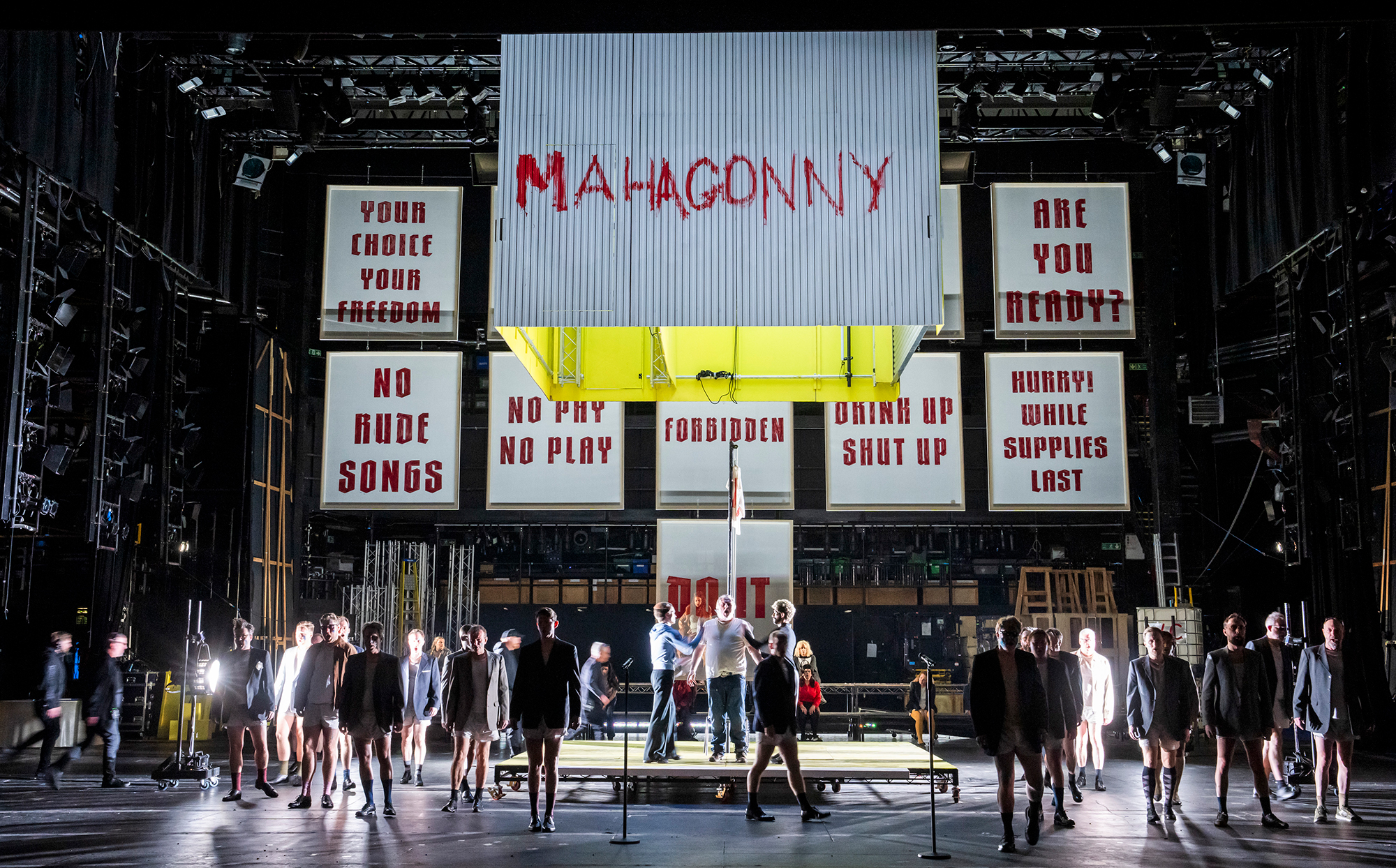 A theatrical stage features actors in modern clothing with bold signs reading phrases like YOUR CHOICE YOUR FREEDOM, FORBIDDEN, and MAHAGONNY in red letters—evoking the dramatic spirit of Rise and Fall in the City of Mahagonny’s industrial backdrop.