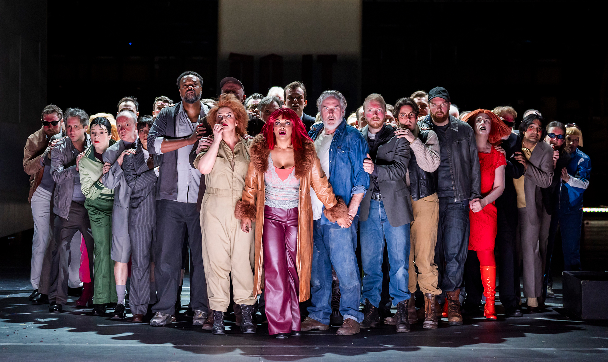 A large group of people in varied costumes stand closely together on stage, some holding each other. Their intense, emotional expressions and dramatic lighting capture the turmoil of the Rise and Fall of the City of Mahagonny opera.