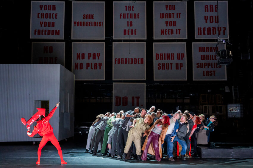 A performer in bright red points upward while a group leans back in unison on stage; behind them, large panels display bold, authoritarian phrases like “THE FUTURE IS HERE,” evoking the striking style of City of Mahagonny.