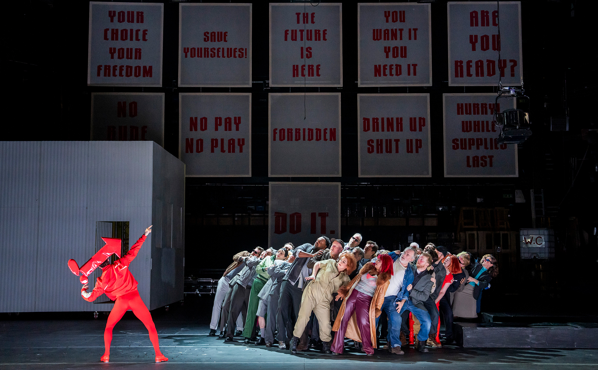 A performer in bright red points upward while a group leans back in unison on stage; behind them, large panels display bold, authoritarian phrases like “THE FUTURE IS HERE,” evoking the striking style of City of Mahagonny.