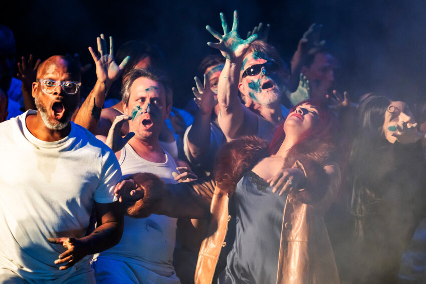 A group of people in dramatic poses, some with green paint on their hands and faces, reach upward and outward amidst smoky lighting, evoking an intense scene from Mahagonny that suggests the rise and fall of a city through powerful performance.