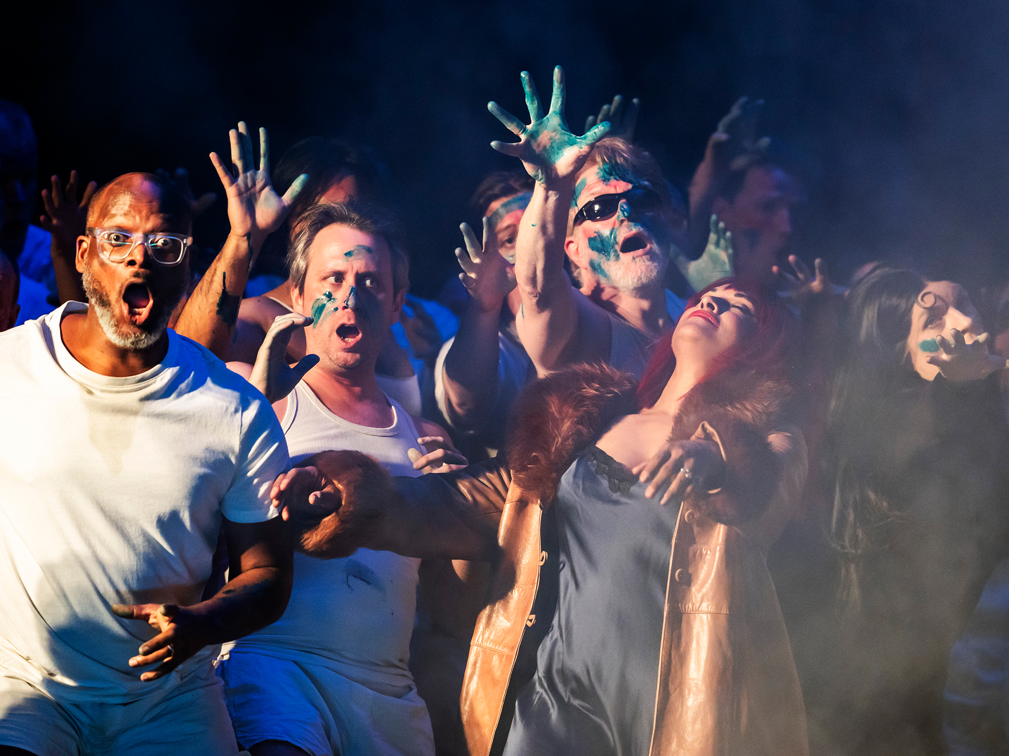 A group of people in dramatic poses, some with green paint on their hands and faces, reach upward and outward amidst smoky lighting, evoking an intense scene from Mahagonny that suggests the rise and fall of a city through powerful performance.