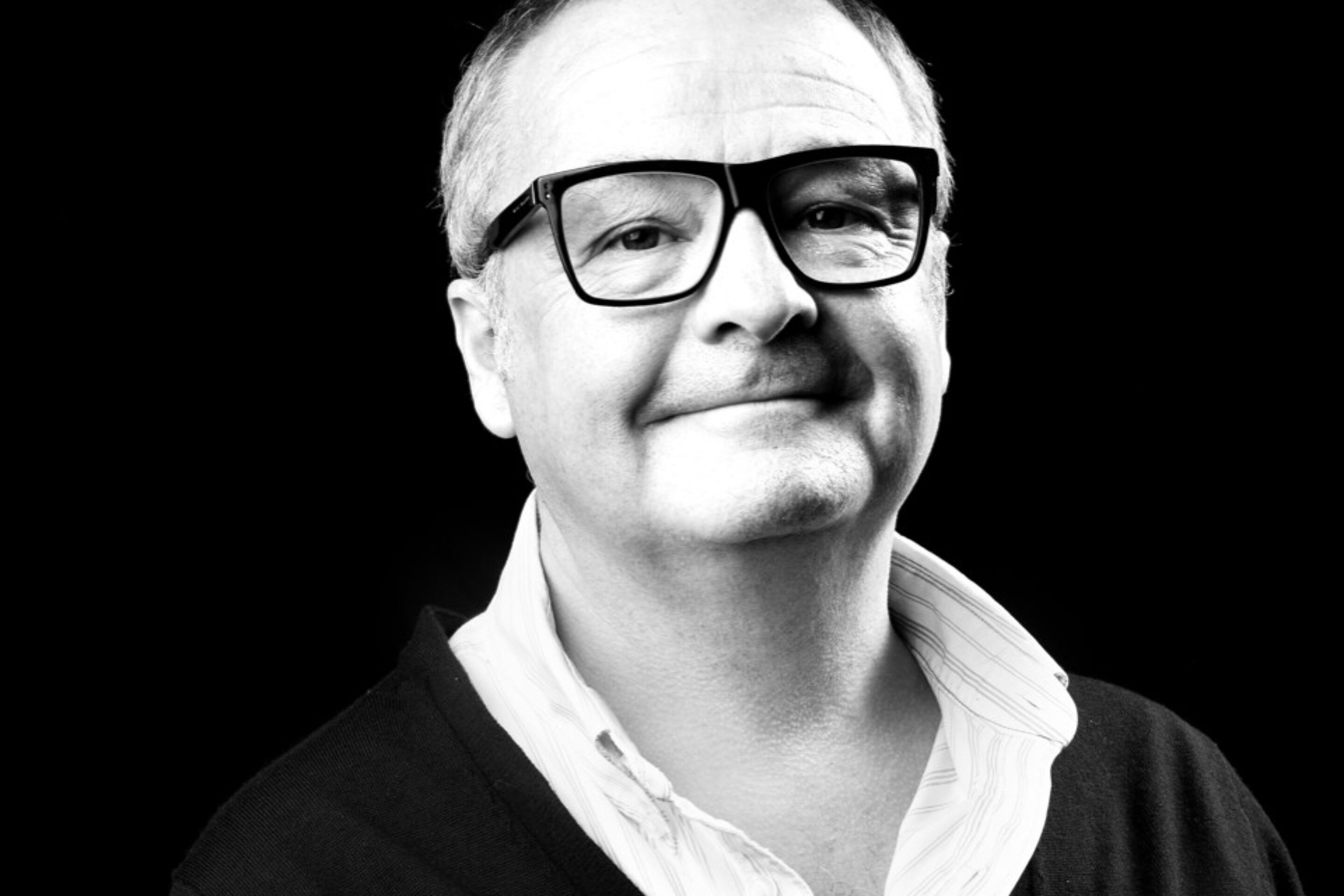 Black and white portrait of Tim Mitchell, a middle-aged man with short hair, wearing large glasses, a white open-collar shirt, and a dark cardigan, smiling slightly against a dark background.
