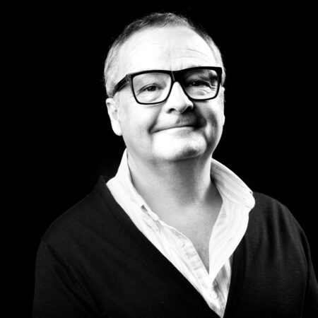 Black and white portrait of Tim Mitchell, a middle-aged man with short hair, wearing large glasses, a white open-collar shirt, and a dark cardigan, smiling slightly against a dark background.