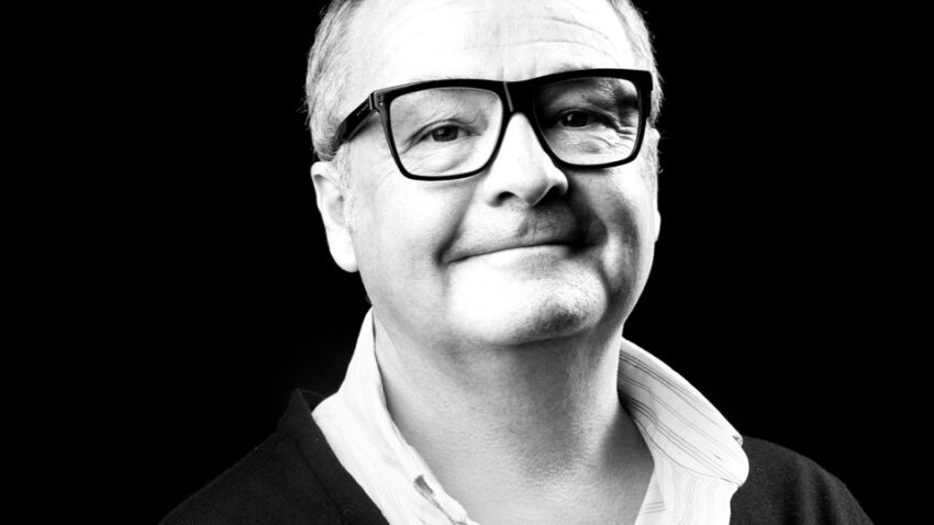 Black and white portrait of Tim Mitchell, a middle-aged man with short hair, wearing large glasses, a white open-collar shirt, and a dark cardigan, smiling slightly against a dark background.