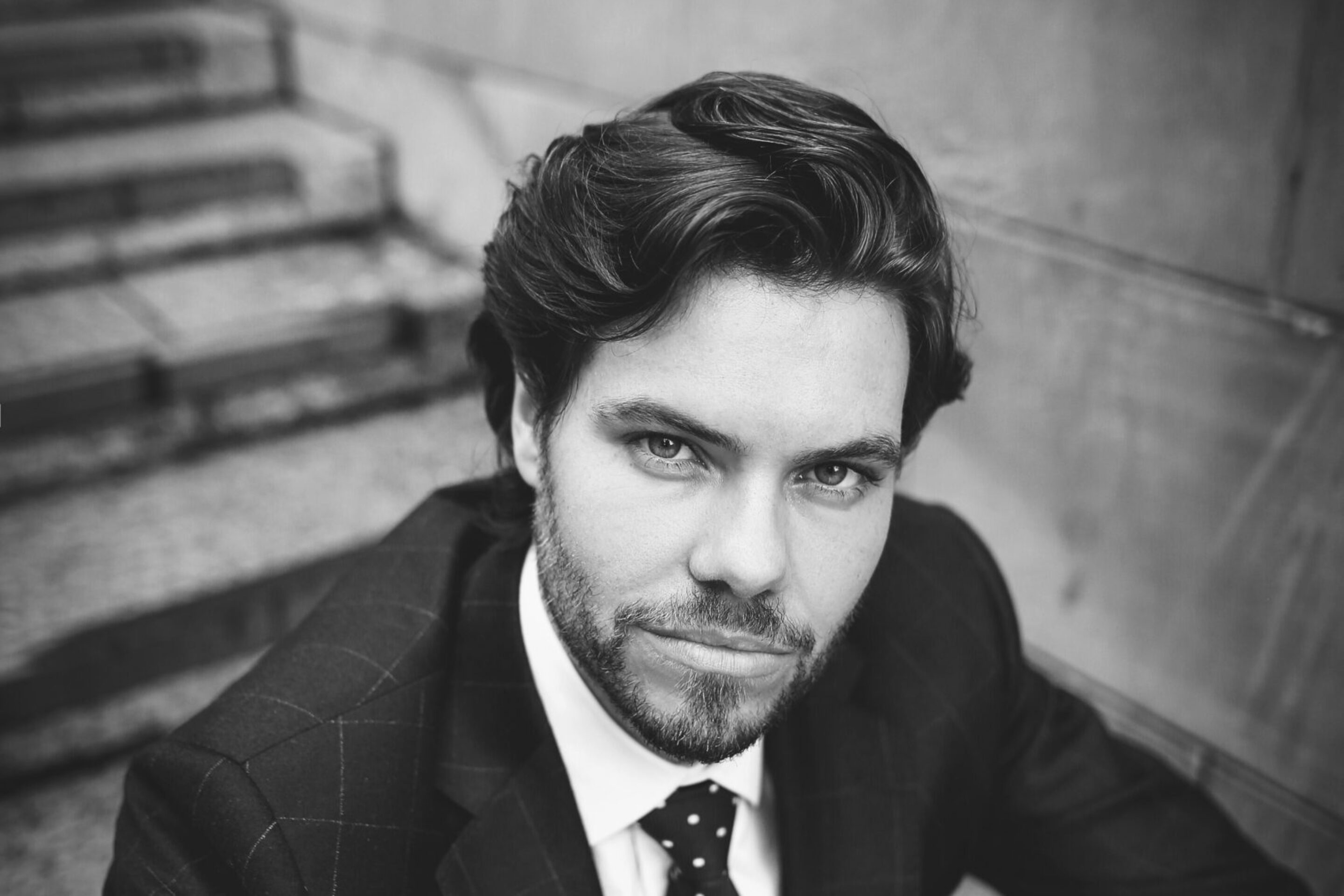 Black and white photo of Thomas Atkins, a man with wavy hair and a beard, wearing a checked suit and spotted tie, sitting on outdoor steps and looking directly at the camera with a serious expression.