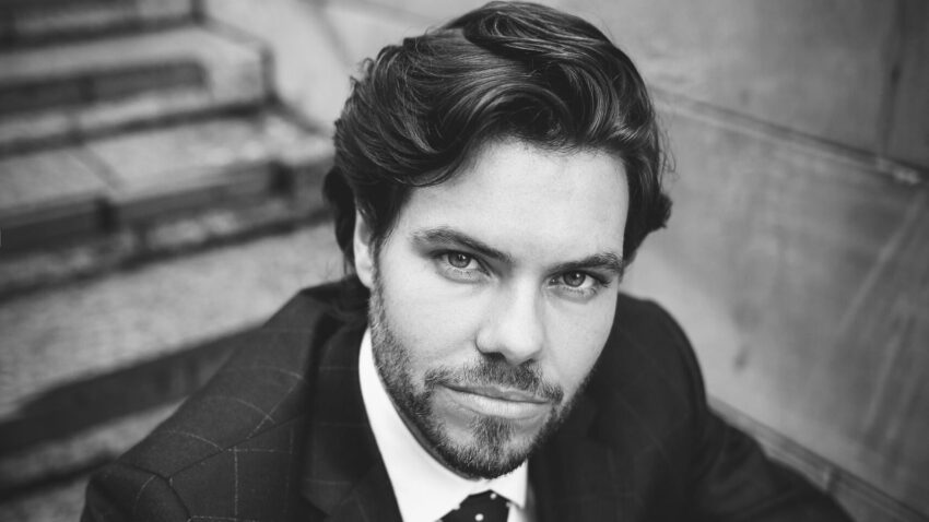 Black and white photo of Thomas Atkins, a man with wavy hair and a beard, wearing a checked suit and spotted tie, sitting on outdoor steps and looking directly at the camera with a serious expression.