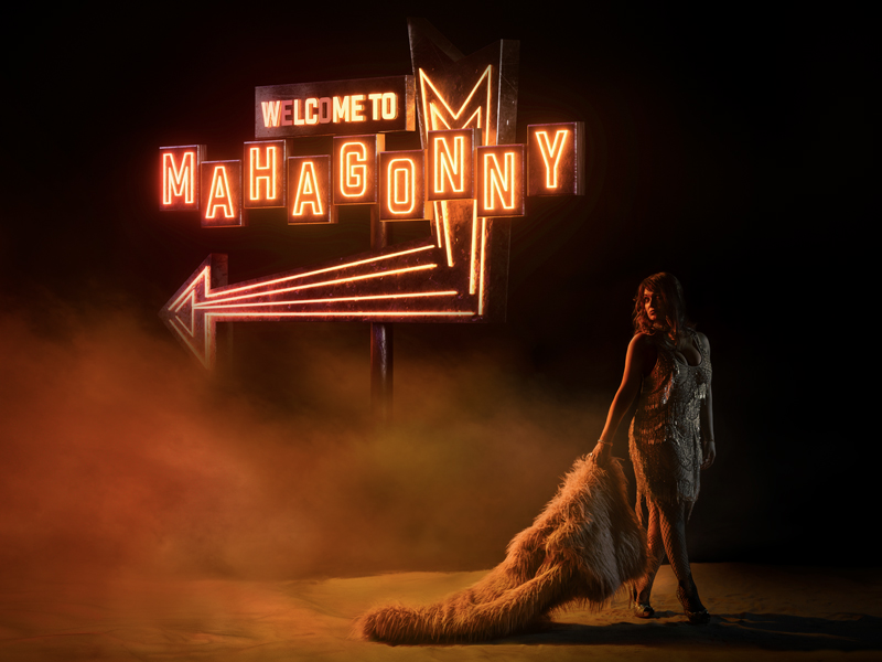 A woman in a sparkling dress stands before a neon sign reading Welcome to Mahagonny, orange light and fog swirling around her. Channelling the spirit of Bertolt Brecht’s Mahagonny opera, she holds a long, fluffy fur coat trailing on the ground.