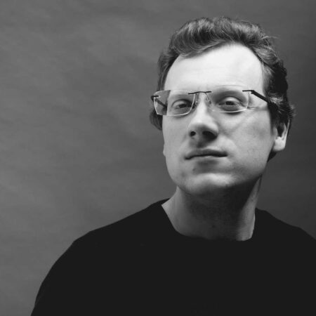 William Cole, wearing spectacles and a dark shirt, looks confidently at the camera against a plain, light-coloured background in this striking black and white portrait.