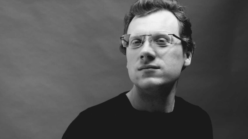 William Cole, wearing spectacles and a dark shirt, looks confidently at the camera against a plain, light-coloured background in this striking black and white portrait.