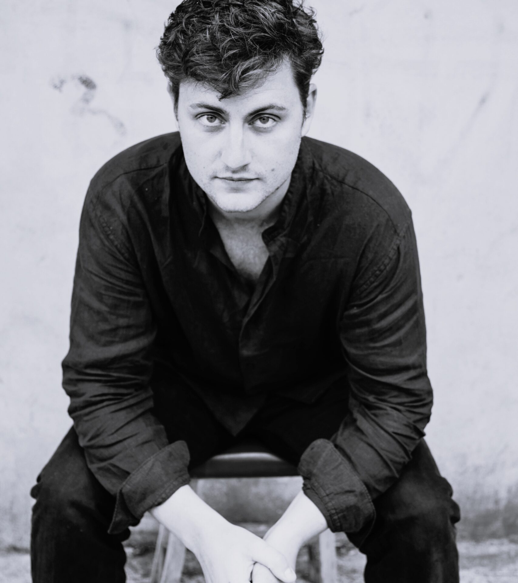 A man with curly hair sits on a stool, leaning forward with his hands clasped. Dressed in a dark shirt and trousers, William Thomas looks directly at the camera with a serious expression against a plain, light-coloured background.