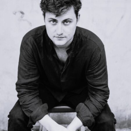 A man with curly hair sits on a stool, leaning forward with his hands clasped. Dressed in a dark shirt and trousers, William Thomas looks directly at the camera with a serious expression against a plain, light-coloured background.