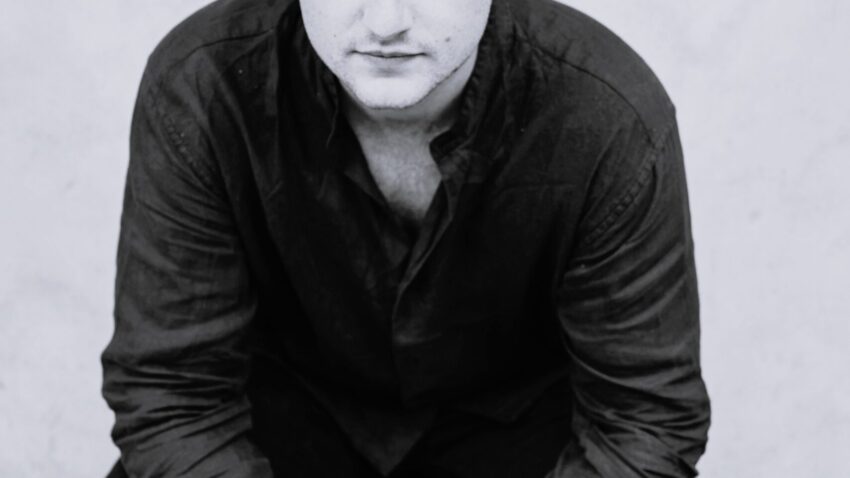 A man with curly hair sits on a stool, leaning forward with his hands clasped. Dressed in a dark shirt and trousers, William Thomas looks directly at the camera with a serious expression against a plain, light-coloured background.