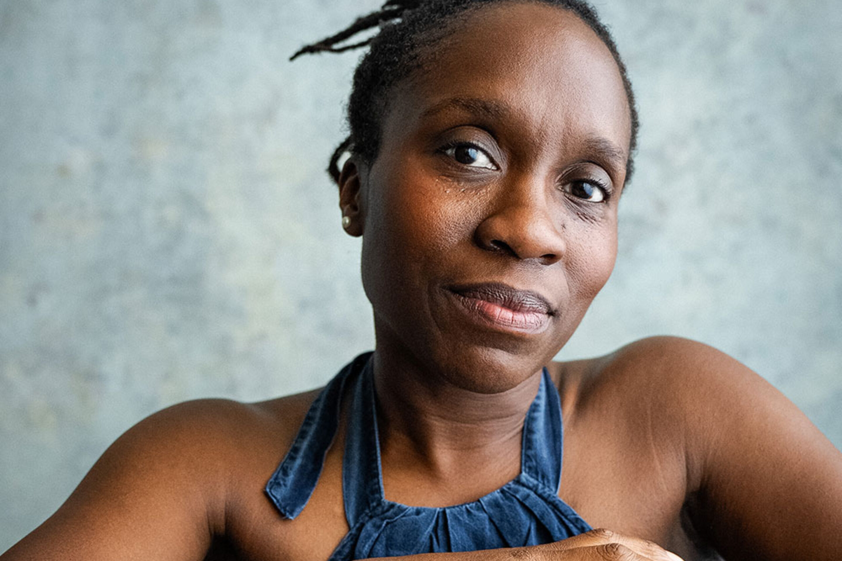 A woman with short, twisted hair sits on a wooden chair, her arms resting on the backrest. Ingrid Mackinnon wears a sleeveless blue top and looks calmly at the camera against a textured, light-coloured background.