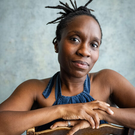 A woman with short, twisted hair sits on a wooden chair, her arms resting on the backrest. Ingrid Mackinnon wears a sleeveless blue top and looks calmly at the camera against a textured, light-coloured background.
