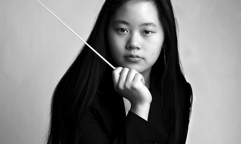 Headshot of Yi-Chen-Lin. Image of a young woman with long dark hair wears a black blazer and holds a conductor’s baton, gazing seriously at the camera against a plain, light background.