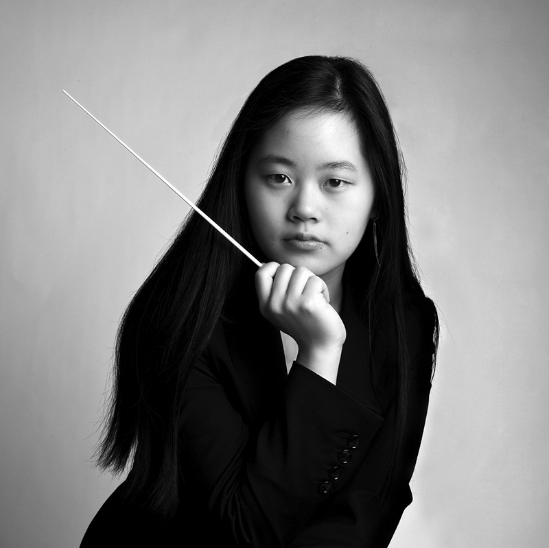 Headshot of Yi-Chen-Lin. Image of a young woman with long dark hair wears a black blazer and holds a conductor’s baton, gazing seriously at the camera against a plain, light background.