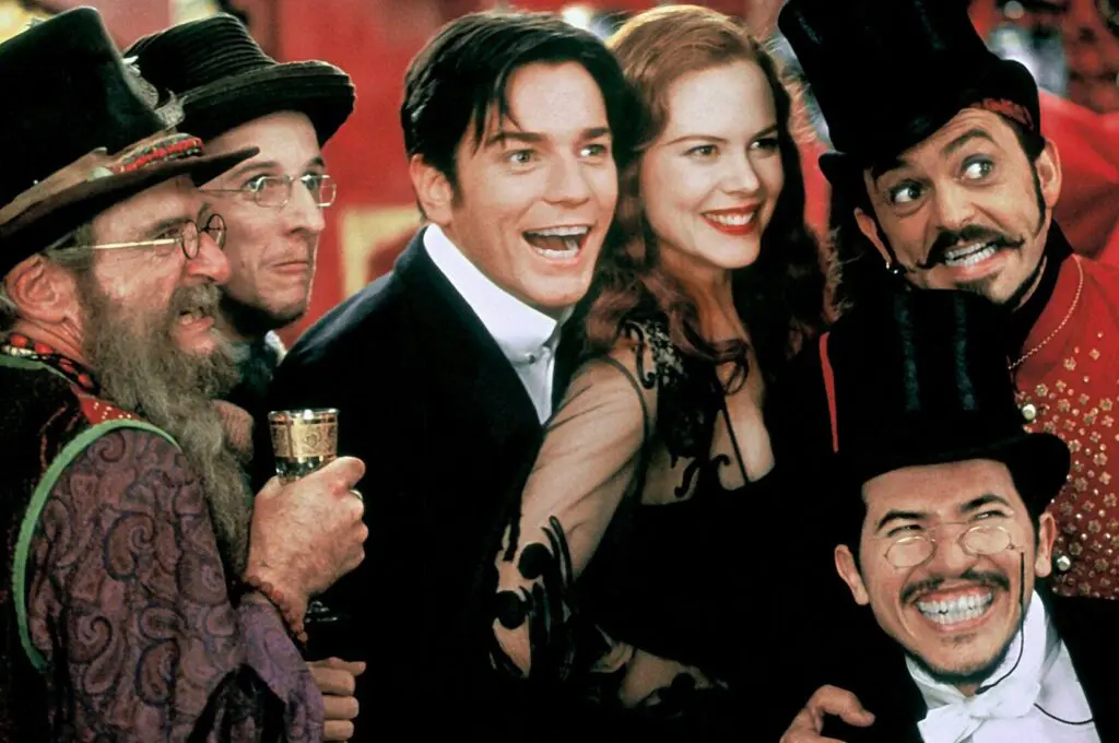 A scene from Moulin Rouge - A group of six people dressed in colourful, theatrical costumes smile and pose together at a lively, festive event with a red background.