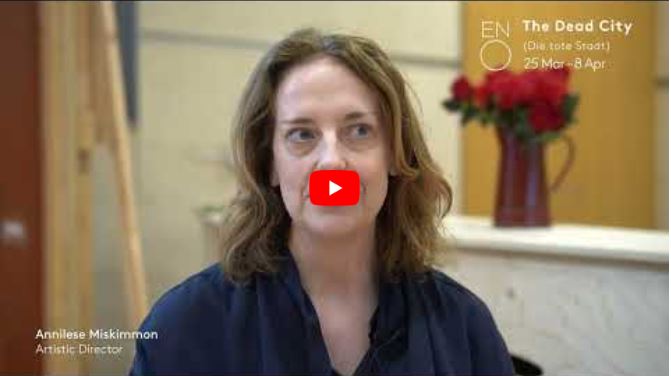 A woman with shoulder-length brown hair sits indoors, facing the camera, giving an introduction. Text on screen reads: “Annelise Miskimmon Artistic Director” and “ENO The Dead City 25 Mar–8 Apr.” A vase of red flowers is blurred in the background.