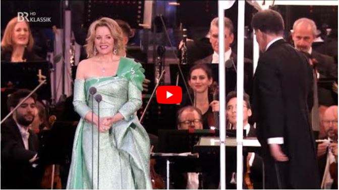 A woman in a sparkling green dress stands on stage smiling, with an orchestra and conductor behind her, as she prepares for the introduction of The Dead City during a classical music performance.