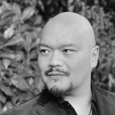 Black and white portrait of Freddie Tong, a bald man with a goatee, wearing a jacket and looking thoughtfully to his left. Blurred foliage is visible in the background.