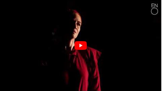 A person in a red outfit stands in dramatic lighting, their face shadowed against a black background.
