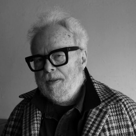 An older man with white hair, a beard, and large black glasses—Herbert Murauer—wears a checked jacket over a collared shirt, looking at the camera against a plain background in black and white.