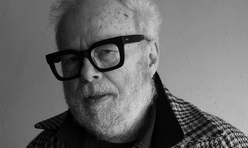 An older man with white hair, a beard, and large black glasses—Herbert Murauer—wears a checked jacket over a collared shirt, looking at the camera against a plain background in black and white.