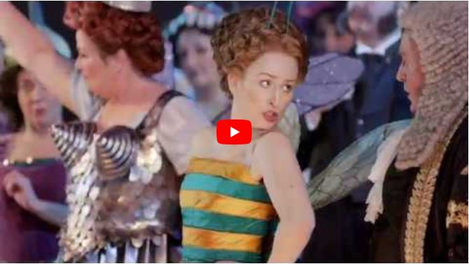 A woman in a striped yellow and green costume with wings and curly hair stands centre stage, surrounded by opera characters in colourful, elaborate outfits at what appears to be a theatrical or operatic performance.