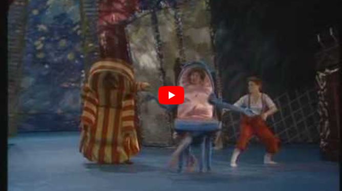 Three people in colourful, whimsical costumes perform on a stage with a blue floor and forest-like background.