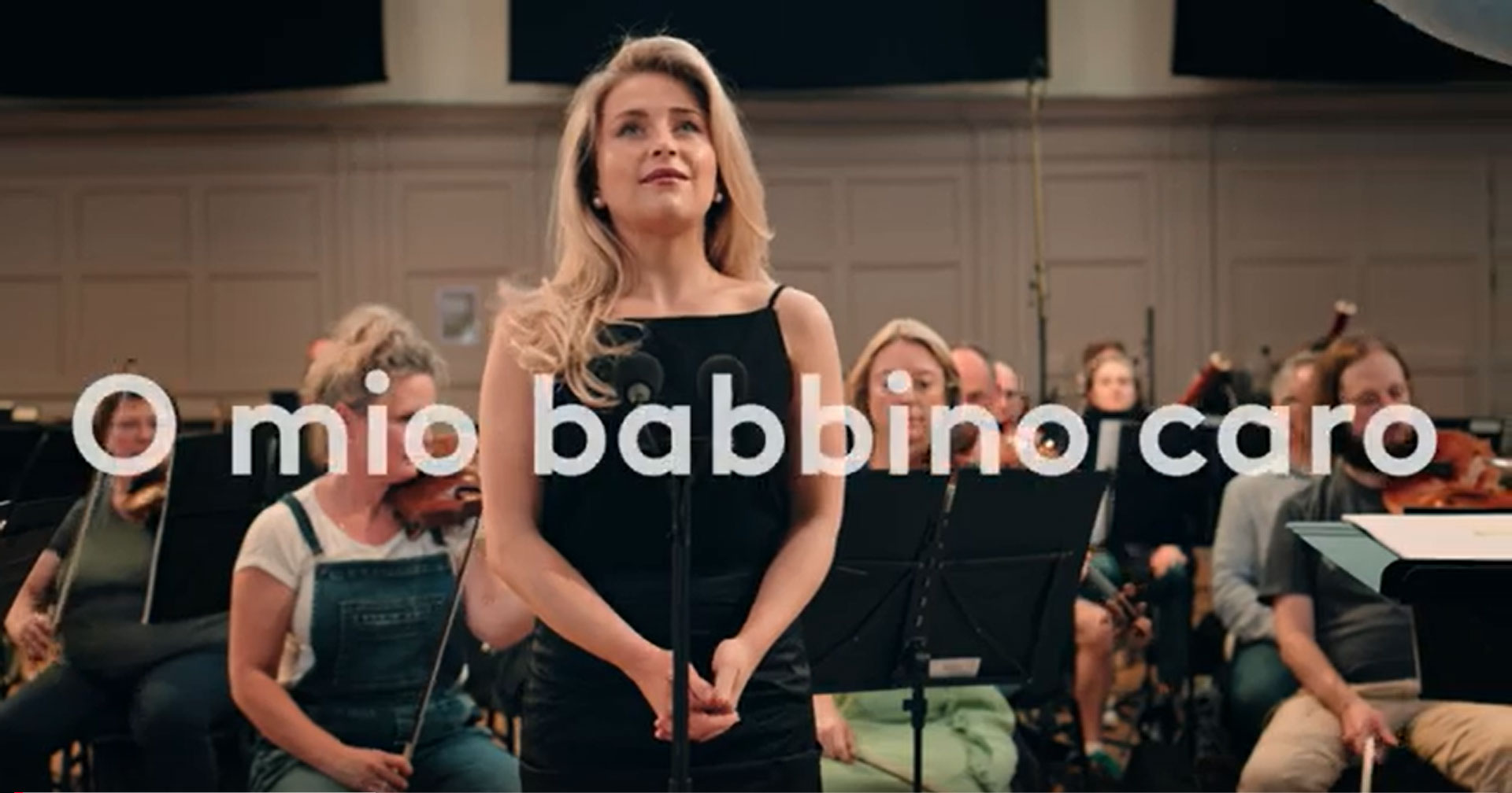 Alexandra Oomens, a woman in a black dress stands at a microphone, singing with an orchestra behind her. Large text across the image reads O mio babbino caro, one of the most famous opera songs. Musicians play string instruments in a concert hall setting.