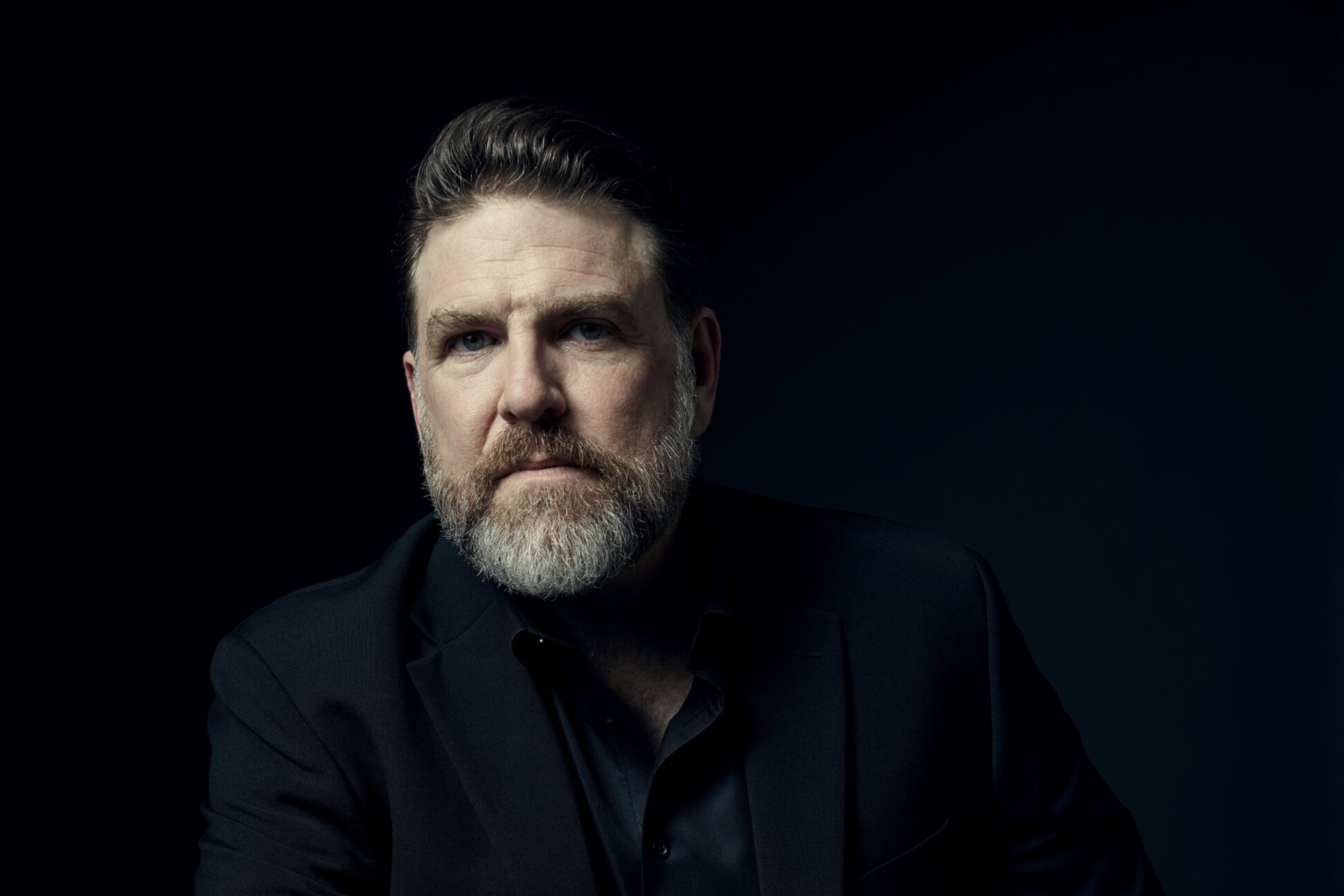 A man with a beard and short hair sits at a table, wearing a black suit and shirt, looking ahead with a serious expression. The dark background highlights Michael Mayes’s face and his hand resting on the table.
