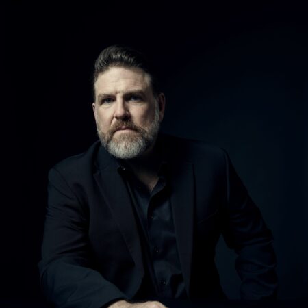 A man with a beard and short hair sits at a table, wearing a black suit and shirt, looking ahead with a serious expression. The dark background highlights Michael Mayes’s face and his hand resting on the table.