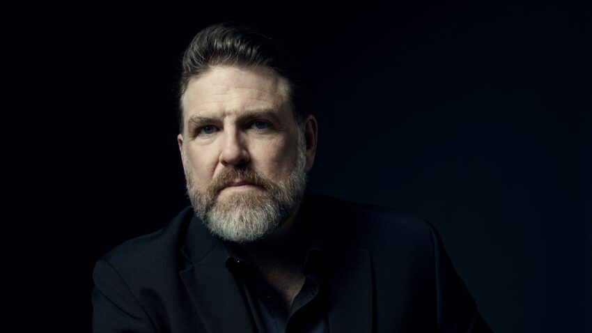 A man with a beard and short hair sits at a table, wearing a black suit and shirt, looking ahead with a serious expression. The dark background highlights Michael Mayes’s face and his hand resting on the table.