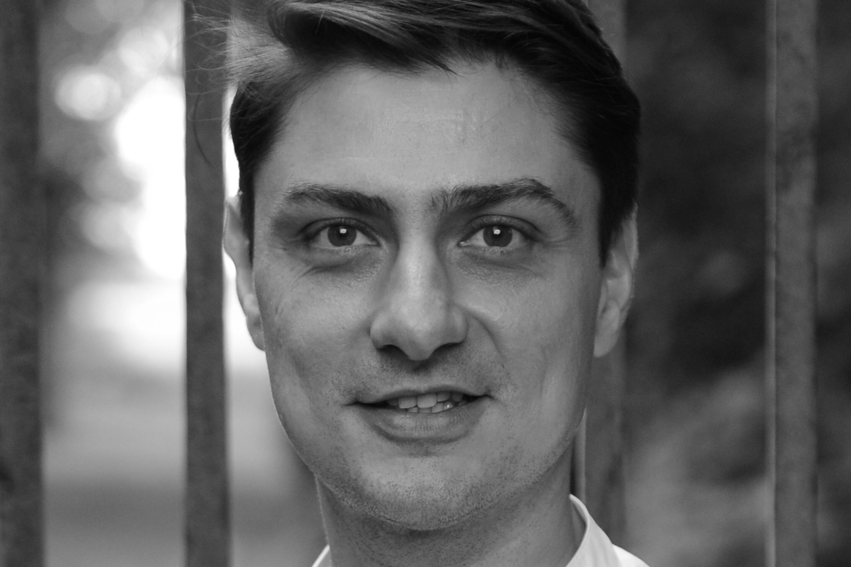 A man with short dark hair, wearing a white collared shirt, stands outdoors in front of vertical metal bars. The background is blurred with greenery, and the photo is in black and white.