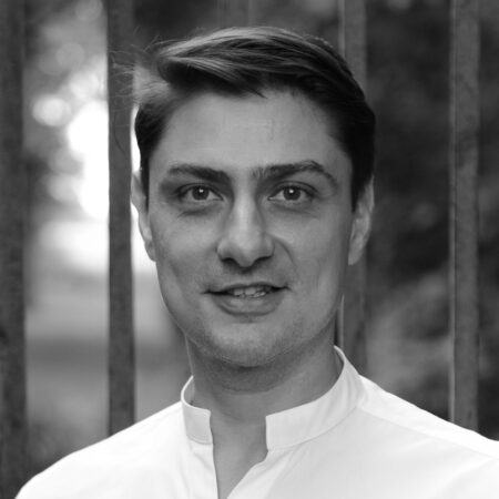 A man with short dark hair, wearing a white collared shirt, stands outdoors in front of vertical metal bars. The background is blurred with greenery, and the photo is in black and white.