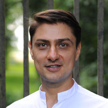 A man with short brown hair wearing a white shirt stands outdoors in front of vertical metal bars and greenery, smiling slightly at the camera.