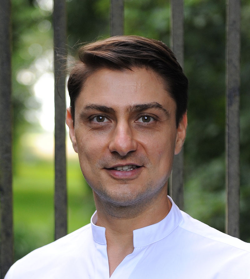 A man with short brown hair wearing a white shirt stands outdoors in front of vertical metal bars and greenery, smiling slightly at the camera.