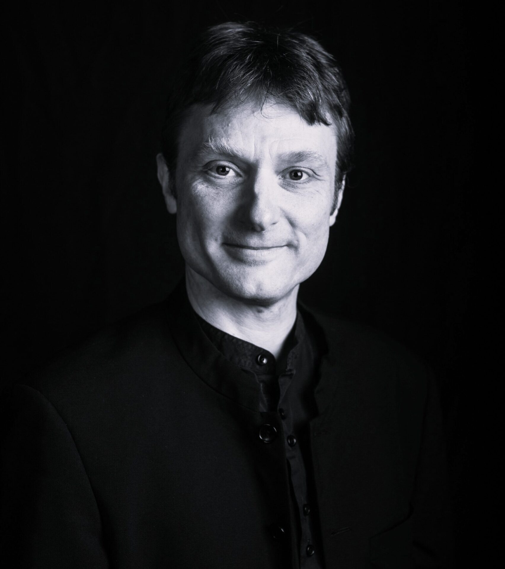 Black and white portrait of a person with short hair, wearing a buttoned-up dark jacket, smiling gently, and looking directly at the camera against a dark background.