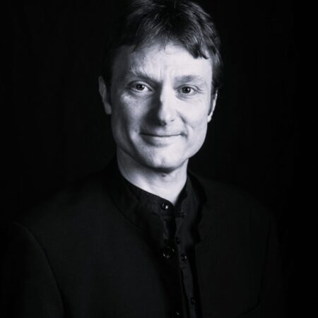 Black and white portrait of a person with short hair, wearing a buttoned-up dark jacket, smiling gently, and looking directly at the camera against a dark background.