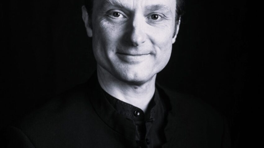 Black and white portrait of a person with short hair, wearing a buttoned-up dark jacket, smiling gently, and looking directly at the camera against a dark background.