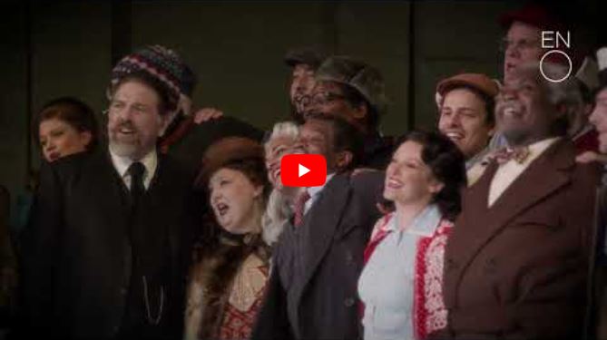 A diverse group of people dressed in vintage winter clothing stand close together, smiling and singing on stage in a scene inspired by It’s a Wonderful Life. A red play button overlays the image, with the ENO logo in the top right corner.
