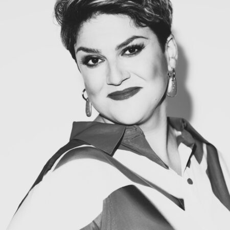 Black-and-white portrait of Rosie Aldridge, a woman with short, styled hair, wearing hoop earrings and a collared shirt with a bold pattern. She is smiling slightly and looking confidently at the camera.