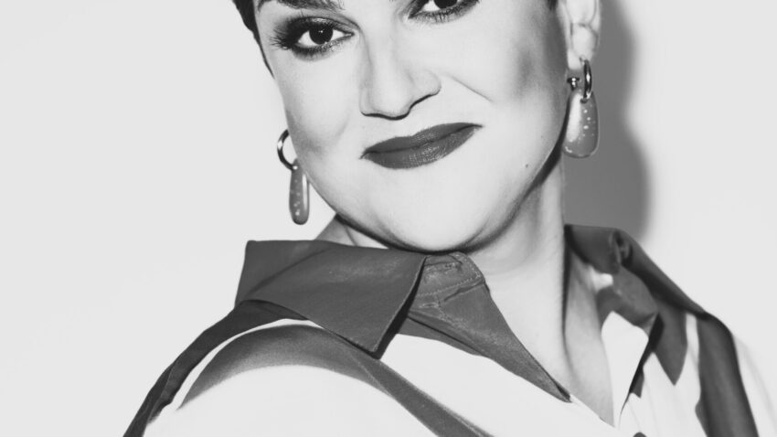 Black-and-white portrait of Rosie Aldridge, a woman with short, styled hair, wearing hoop earrings and a collared shirt with a bold pattern. She is smiling slightly and looking confidently at the camera.