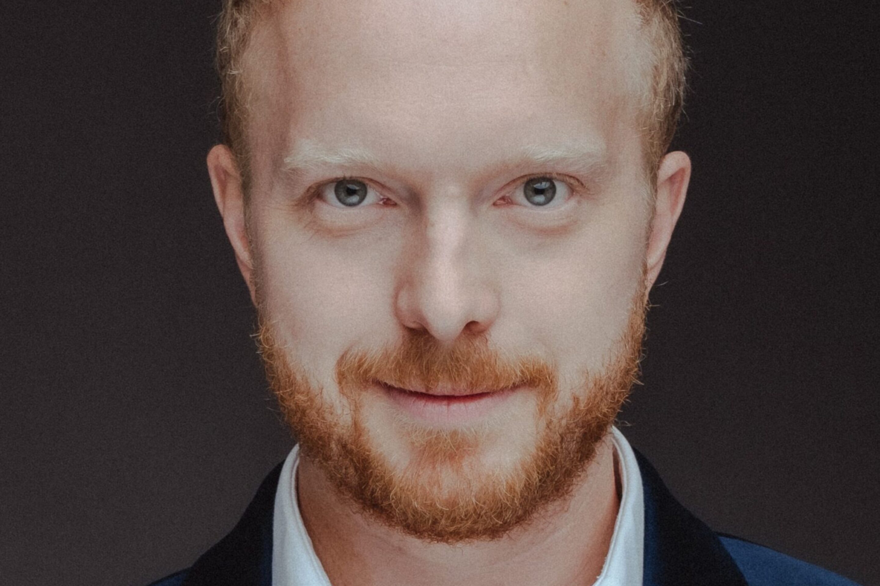 Alex Otterburn, a man with short, curly blond hair and a reddish beard, is wearing a dark blue velvet blazer and a white dress shirt, looking directly at the camera with a neutral expression against a dark background.
