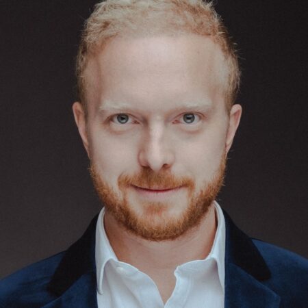 Alex Otterburn, a man with short, curly blond hair and a reddish beard, is wearing a dark blue velvet blazer and a white dress shirt, looking directly at the camera with a neutral expression against a dark background.