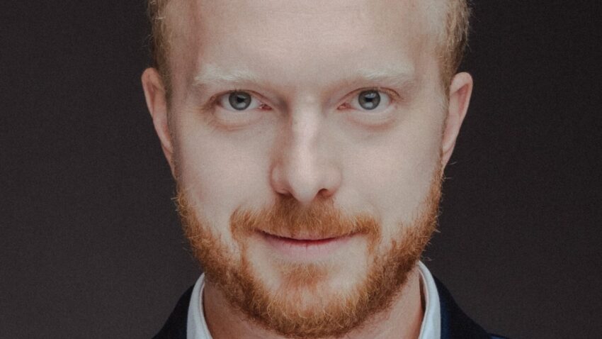 Alex Otterburn, a man with short, curly blond hair and a reddish beard, is wearing a dark blue velvet blazer and a white dress shirt, looking directly at the camera with a neutral expression against a dark background.