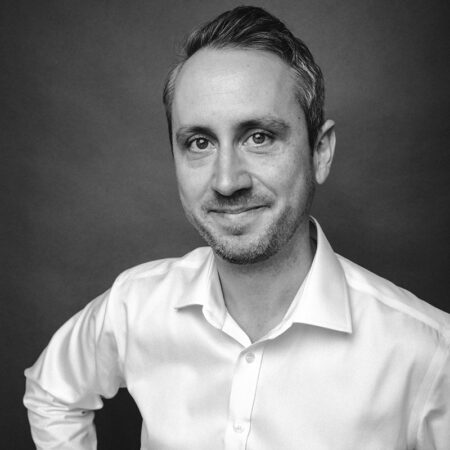 Christopher Nairne, a man with short hair and light stubble, wearing a white button-up shirt, smiles slightly whilst looking at the camera. The photo is in black and white with a plain dark background.